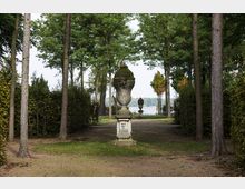 Parklandschaft mit einem gepflasterten Weg, der von hohen Bäumen und Hecken gesäumt ist. In der Mitte steht ein verzierter Steinsockel mit einer großen, bemoosten Steinvase; im Hintergrund ist ein See sichtbar.