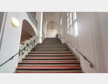 Breite Treppe in einem historischen Gebäude mit kunstvoll verzierten Geländern, hohen weißen Wänden und großen Fenstern, durch die viel Licht fällt. An der Decke hängt eine klassische Laterne, und ein roter Teppich bedeckt die Stufen.