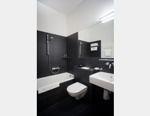 Modernes Badezimmer mit dunklen Fliesenwänden, einer weißen Badewanne mit Duschkopf, einem Hänge-WC und einem rechteckigen Waschbecken. Ein großer Spiegel sowie ein Handtuchhalter mit weißen Handtüchern sind ebenfalls sichtbar.