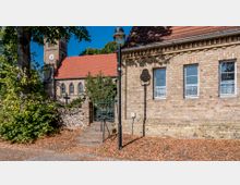 Historische Backsteinkirche mit einem roten Ziegeldach und einem Glockenturm im Hintergrund, umgeben von Bäumen und einer Steinmauer mit einem Eisentor. Im Vordergrund ein Gebäude mit gelbem Ziegelmauerwerk, Fenster und einer Straßenlaterne.
