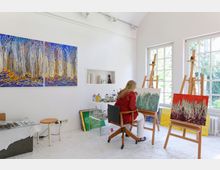 Ein helles Kunstatelier mit großen Fenstern, das von Tageslicht durchflutet wird. Eine Künstlerin sitzt auf einem Stuhl und malt an einem abstrakten Gemälde auf einer Staffelei; um sie herum sind weitere Gemälde, Farben und Künstlerutensilien zu sehen.