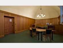 Holzgetäfelter Raum mit gewölbter Decke, zentralem Tisch und mehreren Stühlen mit grünen Polstern. Im Hintergrund befinden sich eine holzverkleidete Tür sowie ein kleiner Arbeitsplatz mit Monitoren und Lampen.