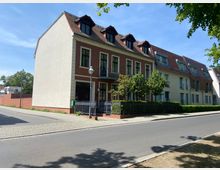 Straßenszene mit einem mehrstöckigen Wohngebäude mit roten Dachgiebeln und hellen Fassaden, das angrenzend an einen Bürgersteig steht. Vor dem Gebäude befinden sich ein kleiner Garten mit Büschen und Bäumen sowie ein Zaun mit einem Eingangstor.