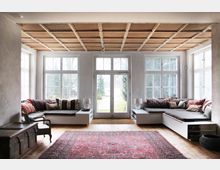 Wohnraum mit großen, weißen Sprossenfenstern und einer Glastür, die zu einem Garten führt. Der Raum hat eine Holzdecke, eingebaute Sitzbänke mit bunten Kissen, einen orientalischen Teppich und helle Wände.