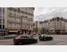 Straßenszene in einer städtischen Umgebung mit mehrstöckigen Wohn- und Geschäftsgebäuden, darunter ein historisches Gebäude mit verzierten Fassadendetails. Im Vordergrund fahren Autos auf der Straße, und entlang der Gebäude befinden sich Geschäfte, darunter ein Café mit der Aufschrift „COFFEE2GO“.