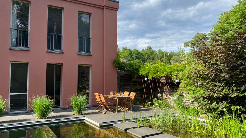 Rückansicht eines modernen, zweigeschossigen Hauses mit rosafarbener Fassade und hohen Fenstern, davor eine Terrasse mit Holztisch und Stühlen. Im Vordergrund liegt ein naturnaher Gartenteich mit Seerosen und Trittplatten, umgeben von dichter Bepflanzung und Sträuchern.