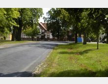 Eine asphaltierte Straße führt durch ein ländliches Dorf mit einem rot-weißen Backsteingebäude im Hintergrund, umgeben von Bäumen und gepflegten Grasflächen. Im Vordergrund sind Schatten von Bäumen sichtbar, während geparkte Autos und weitere Gebäude in der Ferne zu sehen sind.