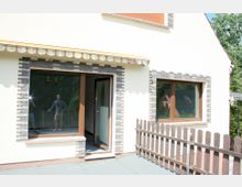 Außenansicht eines Wohnhauses mit weißer Fassade, Fenster- und Türrahmen aus braunem Holz, eingerahmt von dunkelgrauen Klinkersteinen. Im Vordergrund befindet sich ein kleiner Balkon mit Holzgeländer, rechts ist ein Baum sichtbar.