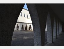 Innenhof eines modernen Gebäudes mit weiß gestrichener Fassade und spitzbogigen Arkaden auf beiden Seiten. Der gepflasterte Boden und der schattige Gang setzen sich in den offenen Innenhof fort.