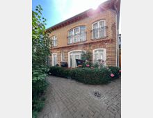 Zweistöckiges Wohnhaus aus gelbem Ziegelstein mit weißen Fensterrahmen und kleinen Balkonen im Obergeschoss. Vor dem Haus befindet sich ein gepflasterter Weg, begrenzt von Sträuchern und Blumen, sowie ein Strandkorb und ein Grill.