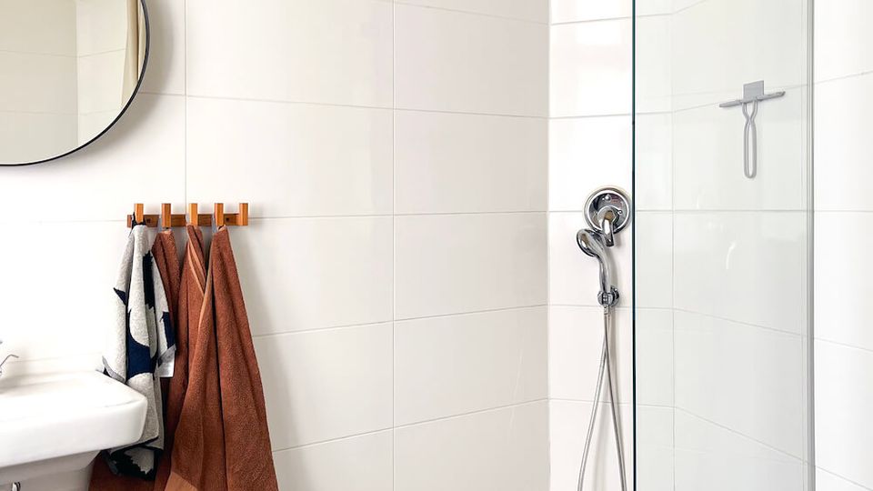Modern bathroom with white tiled walls, a walk-in shower enclosed by a clear glass panel, and a pebble-stone shower floor with a linear drain. A wall-mounted sink, round mirror, and a towel rack with hanging towels sit beside the shower.