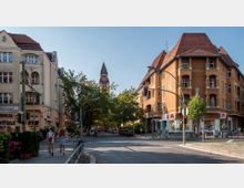 Straßenansicht einer Kreuzung in einer Stadt mit historischen Wohngebäuden und Geschäften. Im Hintergrund ragt ein Kirchturm mit spitzem Dach über Bäume hervor, während im Vordergrund Menschen und Fahrräder sichtbar sind.