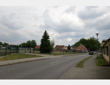 Straßenszene in einem Dorf mit einer asphaltierten Fahrbahn und gepflastertem Gehweg, umgeben von Einfamilienhäusern mit roten Ziegeldächern und einem Drahtzaun. Im Hintergrund stehen vereinzelte Bäume, und der Himmel ist wolkig.