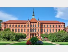 Backsteingebäude im neugotischen Stil mit symmetrischem Aufbau, hohen Rundbogenfenstern, einem zentralen Turm mit Uhr und grüner Spitze. Im Vordergrund ein gepflegter Garten mit bunten Blumenbeeten und Rasenflächen.