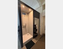 Modernes Badezimmer mit offener Duschkabine in schwarzer Rahmenkonstruktion, weißer Duschtasse und wandmontierter Armatur. Daneben ein kompakter Waschtischbereich mit Spiegel, schwarze Ablagen und eine weiße Ziegelwand unter einer gewölbten Decke.