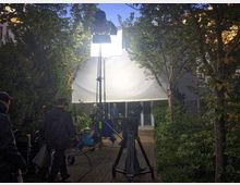 Eine Filmszene wird im Freien gedreht, mit einer großen weißen Diffusorleinwand, die zwischen Bäumen aufgespannt ist. Im Vordergrund befinden sich Kameras auf Stativen, Beleuchtungsausrüstung und Crewmitglieder, umgeben von dichtem Grün und einem gepflasterten Weg.