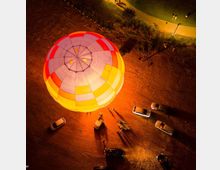 Luftaufnahme eines beleuchteten Heißluftballons auf einer offenen, erdigen Fläche, umgeben von mehreren geparkten Autos und einigen Menschen. Im Hintergrund ist ein beleuchteter Gehweg mit Grünflächen zu sehen.