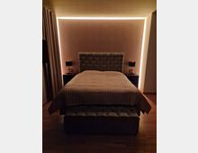 Ein modernes Schlafzimmer mit einem gepolsterten Doppelbett in der Mitte, flankiert von zwei Nachttischen mit schwarzen Lampen. Die Wand hinter dem Bett ist mit indirekter, warmweißer Beleuchtung akzentuiert, und der Raum hat einen Holzboden sowie geschlossene Vorhänge auf der linken Seite.