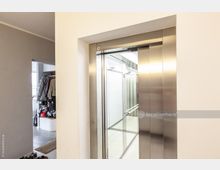 Moderner Innenraum mit einem offenen Aufzug mit Edelstahlrahmen und Spiegelwänden. Links im Bild führt ein Durchgang zu einem Bereich mit einer Garderobe und Schuhen.