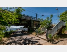 Brücke mit Eisenbahnlinie über einem Weg am Wasser, umgeben von Bäumen und Graffiti an den Betonpfeilern; rechts führt eine Treppe mit Metallgeländer nach oben.