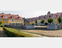 Gelbe, zweigeschossige Reihenhäuser mit roten Satteldächern und kleinen Gauben, angeordnet um einen offenen Platz mit trockenem Rasen, einer niedrigen Steinmauer und wenigen Einzelbäumen. Im Vordergrund befinden sich eine Hecke und eine Treppe, die auf den Platz führen.