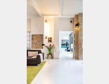 Ein moderner Wohnraum mit weißem Boden, einer schwarzen Couch auf einem grünen Teppich, Dekoration in Form von Wandgemälden, einer Stehlampe und einem Beistelltisch mit einer Blumenvase. Im Hintergrund befinden sich ein heller Durchgang, ein freistehender Backsteinpfeiler und große Fenster, die einen zweiten Bereich sichtbar machen.