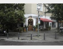 Straßenecke mit einem Café-Restaurant namens "Biscotti", erkennbar an einem Holzportal und einer Beschriftung über dem Eingang. Umgeben von Bäumen, Straßenschildern und kleinen Tischen im Außenbereich; ein rotes Vordach rechts deutet auf ein benachbartes Geschäft oder Café hin.