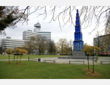 Ein moderner Park mit einer grünen Rasenfläche, kleinen Bäumen und Sitzbänken im Vordergrund. Im Hintergrund befinden sich ein markanter, blauer, turmartiger Kunstbau und ein weiß-graues Bürogebäude mit mehreren Etagen.