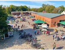 Marktplatz mit Kopfsteinpflaster, auf dem ein Markt mit Ständen, Verkaufswagen und Sitzbereichen stattfindet. Im Hintergrund sind historische Backsteingebäude und Bäume sichtbar, während Fahrräder im Vordergrund abgestellt sind.