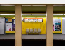 U-Bahn-Station mit gelb gestrichenen Wänden, Werbeplakaten und dem Schild "Bismarckstraße" der Linie U2. Im Vordergrund befinden sich vier metallene Sitzplätze, eingerahmt von gelben Trägern.