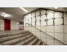 Treppenaufgang in einer U-Bahn-Station mit verschmutzten, beige gefliesten Wänden. Eine Wand zeigt Schäden mit freiliegender Bausubstanz, daneben eine rote Tür und Werbeposter im Hintergrund.