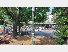 Städtische Straßenszene mit Bäumen und einer Bank im Vordergrund. Im Hintergrund befinden sich eine belebte Straße mit Autos, geparkte Fahrräder, Fußgänger sowie eine erhöhte U-Bahn-Brücke mit Stahlkonstruktion.