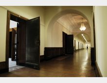 Flur in einem historischen Gebäude mit hohen Decken und großen Rundbögen, links offenstehende dunkle Holztüren, rechts Holzvertäfelung an den Wänden, ein Kronleuchter und Lampen an der Decke; im Hintergrund eine Person am Ende des Flurs.