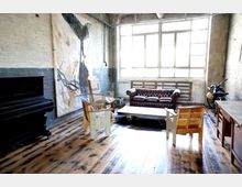 Industrie-inspirierter Raum mit hellen Backsteinwänden, großen Fensterfronten und einem Holzboden. Der Raum ist mit einem dunklen Klavier, einer braunen Chesterfield-Couch, Holzmöbeln und einer minimalistischen Wandgestaltung ausgestattet.