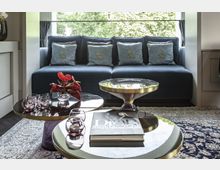 Wohnzimmer mit einem dunkelblauen Sofa, das mit dekorativen Kissen geschmückt ist, im Hintergrund ein großes Fenster mit Blick auf grüne Bäume. Im Vordergrund befinden sich zwei runde Couchtische mit glänzenden Oberflächen, darauf eine Vase mit roten Blumen, kleine Gläser und ein Bildband. Ein gemusterter Teppich bedeckt den Boden.