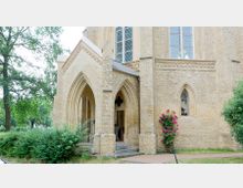 Außenansicht eines historischen Kirchengebäudes aus gelbem Backstein mit spitzbogigem Eingang und Buntglasfenstern. Neben dem Eingang steht ein Rosenstrauch, und im Hintergrund befinden sich Bäume und Grünflächen.