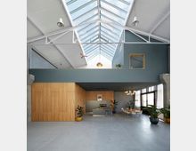 Heller, moderner Innenraum in einem Loftstil mit einer großen Glasdachkonstruktion, die viel Tageslicht hereinlässt. Der Raum ist aufgeteilt in eine Holzverkleidete Wand mit integrierter Kaffeetheke, Sitzbereich mit Tischen und Stühlen sowie Dekoration durch mehrere Zimmerpflanzen.