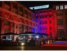 Industriegebäude aus Backstein mit großen Fenstern, nachts beleuchtet in roten und blauen Farben. Im Vordergrund eine Außenterrasse mit Sonnenschirmen und Dekoration; im Hintergrund ein Stahlgerüst, das die Gebäude verbindet.