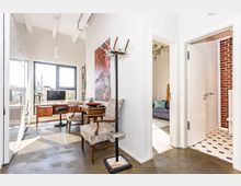 Helle Innenansicht eines modernen Wohnraums mit Betonboden, hohen Decken und großen Fenstern. Sichtbar sind ein Schreibtisch mit Computer, zwei gepolsterte Stühle, ein Kunstwerk an der Wand sowie Zugänge zu einem Wohnzimmer mit grauem Sofa und zu einem Badezimmer mit gemustertem Fliesenboden und roter Backsteinwand.