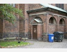 Backsteinfassade eines historischen Gebäudes mit Rundbogenfenstern, einer Tür mit Graffiti und einem kleinen Vordach. Im Vordergrund steht ein Fahrrad neben einer bepflanzten Fläche, während ein blauer Papiercontainer und eine schwarze Mülltonne rechts daneben auf dem gepflasterten Boden zu sehen sind.