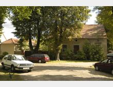 Ein alter, verputzter Bau mit Satteldach und verwitterter Fassade steht hinter Bäumen und Büschen. Im Vordergrund sind mehrere parkende Autos auf einem geschotterten Platz zu sehen.