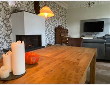 Gemütliches Wohnzimmer mit einem Esstisch aus Holz im Vordergrund, dekoriert mit weißen Kerzen und einem Kürbis. Im Hintergrund sind ein moderner Kamin, eine gemusterte Tapete, ein antikes Klavier, eine graue Couch und ein an der Wand montierter Fernseher zu sehen.