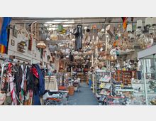 Innenansicht einer großen, hallenartigen Secondhand- und Trödlerladenfläche mit hohen Decken, dicht bestückten Regalen und Vitrinen. Von der Decke hängen zahlreiche Lampen, Haushaltsgegenstände und Dekorationen; entlang eines Mittelgangs stehen Kleiderständer, Einkaufswagen und Warenstände.