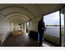 Holzverkleideter Raum in einem kleinen, bogenförmigen Gebäude mit einem Esstisch, Stühlen und einem Bett im Hintergrund. Ein Mann steht an einer großen Glasfront, die Ausblick auf einen Fluss und eine flache Uferlandschaft bietet, während er eine Angelrute hält.