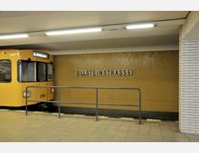 Gelbe U-Bahn im Bahnhof Ullsteinstraße mit einer Wand aus gelben Fliesen und einem Schild mit dem Stationsnamen in schwarzen Großbuchstaben; im Vordergrund ein Geländer und ein Fliesenboden.