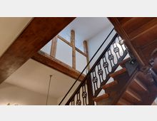 Innenansicht eines Treppenhauses mit einer Holztreppe, einem verzierten Geländer aus dunklem Metall und sichtbaren Holzbalken im oberen Bereich der Wand. Die Wände sind weiß gestrichen, und eine einzelne Pendelleuchte hängt von der Decke.