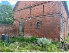 Backsteinfassade eines alten Gebäudes mit einem Holzhoftor und einem kleinen vergitterten Fenster. Im Vordergrund ein überwucherter Gartenbereich mit einer Regentonne und Steinumrandungen, daneben Mauerreste und ein Fallrohr.