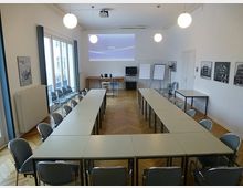 Konferenzraum mit U-förmig angeordneten Tischen und Stühlen, einem Parkettboden, einer Leinwand an der Wand, mehreren Whiteboards, einem Rednerpult und dekorativen Bildern an den Wänden. Drei Hängelampen beleuchten den Raum, der über große Fenster Tageslicht erhält.