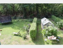 Großer Garten mit Rasenfläche, einem Glashaus auf der rechten Seite und einer geschnittenen Hecke. Links steht ein Trampolin und eine Gartenliege, im Vordergrund ein Tisch mit Töpferwaren und zwei Stühlen. Im Hintergrund ist ein dichter Wald erkennbar.