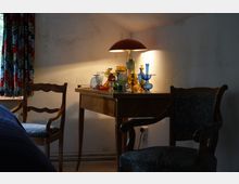 Ein gemütlicher Raum mit einem Holztisch, auf dem verschiedene bunte Glasgefäße stehen, beleuchtet von einer Lampe mit rotbraunem Schirm. Im Vordergrund stehen zwei gepolsterte Holzstühle, im Hintergrund sind bunte Vorhänge zu sehen.